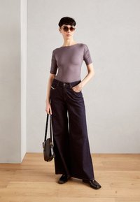 Přiléhavý mauve top s detailem řasení, spárovaný s volnými tmavými denimovými džíny. Doplňky zahrnují velké sluneční brýle a malou černou kabelku.