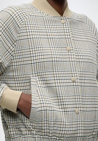 Καρό σακάκι με βάση μπεζ και μπλε και κίτρινες γραμμές, μπεζ ριμπ λαιμόκοψη και μανσέτες, και μπροστινές τσέπες με κουμπιά.