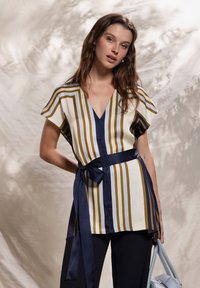 Top a righe in crema, oro e blu navy, caratterizzato da scollo a V, maniche corte e cintura annodata in vita. Abbinato a pantaloni neri e una borsa azzurro chiaro.
