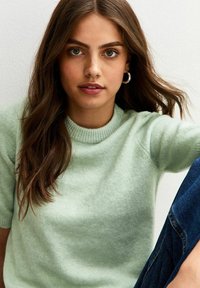 Maglione a maglia verde chiaro, con maniche corte e collo tondo a coste. Il modello ha capelli ondulati e orecchini a cerchio argentati. Sono visibili jeans in denim.