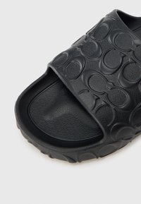 Schwarze Slidesandale mit einer strukturierten Gummi-Oberfläche mit kreisförmigem Muster, konturiertem Fußbett und leicht erhöhtem Absatz.