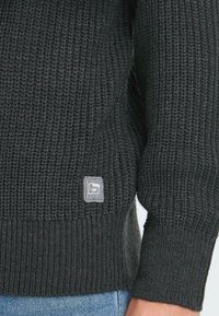 Ciemny, teksturowany sweter z dzianiny z prążkowanym dołem i mankietami. Posiada małą, kwadratową szaro-skalną naszywkę z logo w dolnym prawym rogu. ✨