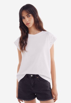 T-shirt en coton blanc avec des manches courtes ornées de perforations. Accompagné de shorts en jean noir avec un ourlet effiloché.