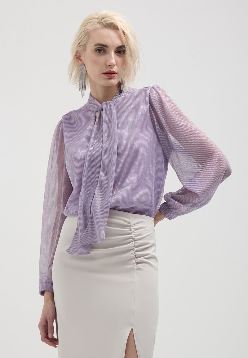 Blouse lilas à rayures verticales, manches longues transparentes, détail à nouer au cou, assortie à une jupe crème ajustée avec fronces sur le côté et une fente.