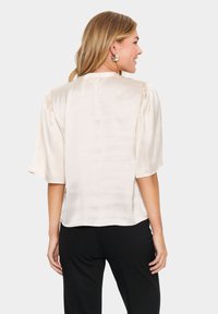 Blusa in seta a maniche corte color crema con una texture liscia e lucida. Presenta leggere pieghe sulle spalle e un collo rotondo.