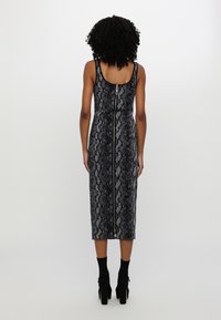 Femme aux cheveux bouclés portant une robe sans manches, mi-longue, noire et grise à motif peau de serpent avec une fermeture éclair dans le dos et des bottines noires à talons hauts.