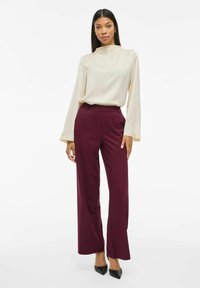 Blusa di seta crema con maniche lunghe e collo alto abbinata a pantaloni larghi bordeaux e tacchi a punta neri.