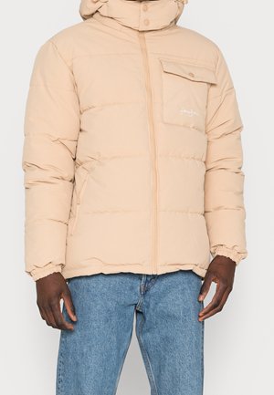 Veste matelassée beige avec fermeture éclair à l'avant, poche poitrine à bouton, poignets élastiques, portée avec un jean bleu, vue du cou jusqu'aux hanches.