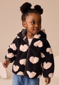 Veste en polaire noire avec de grands motifs de cœur beiges, fermeture éclair à l'avant et capuche. Texture douce, associée à une chemise légère et un jean en denim.