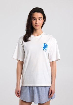 Woman with dark hair wearing a white oversized t-shirt with a small blue abstract print on the chest and a blue-striped skirt.