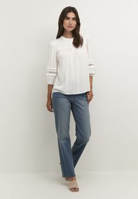 Blusa bianca con pieghe verticali e scollo rotondo, caratterizzata da maniche a sbuffo a tre quarti rifinite in pizzo. Abbinata a jeans a gamba dritta blu chiaro.