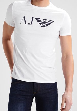 Vit bomulls t-shirt med en svart logotyp som har stiliserade bokstäverna "AJ" och "GA" tillsammans med vingmotiv. Kort ärm och rundad halsringning.