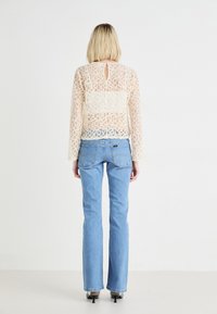 Top de encaje beige transparente con patrón circular, mangas largas y detalle en ojal en la parte posterior, combinado con jeans de campana azul claro y tacones negros.