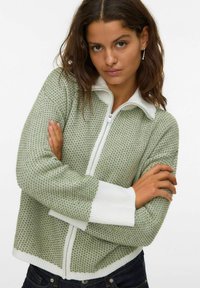 Zielony sweter na zamek z dzianiny, z białymi prążkowanymi mankietami i kołnierzykiem, wyróżniający się teksturowanym wzorem i skróconym fasonem.