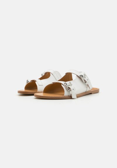 Sandale din piele albă cu un design deschis la vârf, accente cu cataramă ajustabilă și o talpă flată maro. Textură netedă și stil minimalist.