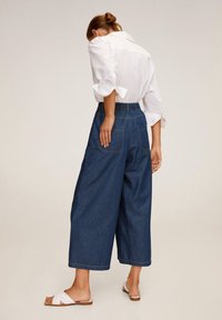 Culottes en denim à taille haute et jambes larges, dotées de poches arrière et d'une coupe décontractée. Associés à une chemise blanche à boutons et des sandales.