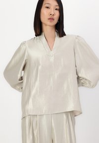 Blusa argentata chiara con scollo a V, maniche a palloncino e una texture liscia, leggermente lucida. Presenta una vestibilità ampia e un dettaglio di lucentezza sottile.