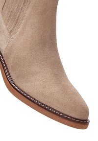 Beige mokkasiini nilkkasaappaat, joissa on terävä kärki, nahkapohja ja kontrastiväriset tikkaukset reunoilla. Ribatuille sivupaneeleille joustavuuden lisäämiseksi.