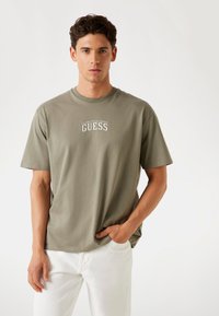 Olivově zelené bavlněné tričko s krátkými rukávy, kulatým výstřihem a bílým textovým logem "GUESS" na hrudi. Kombinováno s bílými džíny.