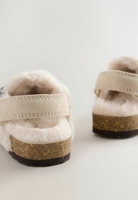 Heledadbeige klotsid pehme ja kiharate sisepindadega ning korkjalaga. Omab reguleeritavat rihma ja tekstureeritud kummist väljalappi.