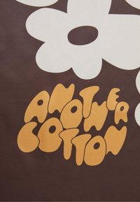 Brun baggrund med store hvide blomster. Teksten er skrevet med legeskrift i rund, orange skrifttype og siger "ANOTHER COTTON." Glat tekstur.