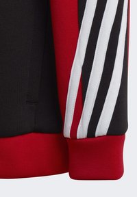 Black and red sweatshirt sleeve with three white stripes, made of textured fabric. Features a solid red cuff at the bottom.