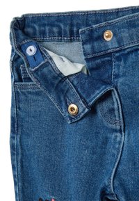 Jeans de mezclilla con un lavado azul claro. Presenta herrajes de latón, un cierre de botón y detalles de costura. Pequeño diseño bordado en la parte frontal.