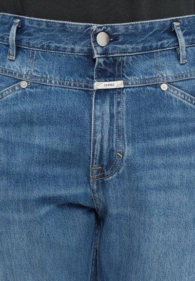 Blugi din denim albastru, văzuți din față, cu închidere cu buton metalic, bucle pentru curea și o etichetă mică albă care spune "CLOSED" deasupra zonei fermoarului.