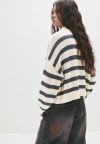 Strickpullover mit horizontalen marineblauen Streifen auf einem cremefarbenen Hintergrund, mit einem verkürzten Design und geripptem Saum. Kombiniert mit dunklen Jeans, die ein rundes Logo tragen.