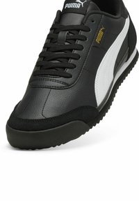 Sneaker PUMA noir et blanc avec une tige en cuir texturé, des lacets noirs et une semelle en caoutchouc. Présente un logo PUMA doré sur le côté.