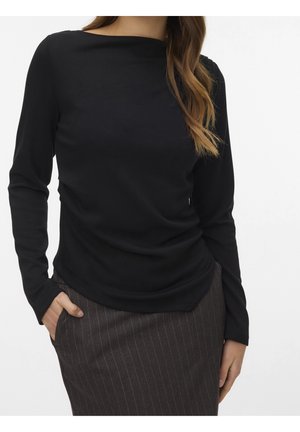 Femme portant un haut noir moulant à manches longues et une jupe à fines rayures gris foncé, avec une main dans la poche de la jupe.