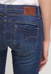 Denim jeans in dark blue with a smooth texture. Features a leather label at the waistband and curved stitching on the back pockets.