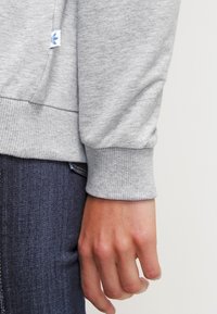 Nära bild av en hand bredvid en grå sweatshirtärm med ribbad mudd och blå jeans, som visar tygtextur och en liten klädlapp.
