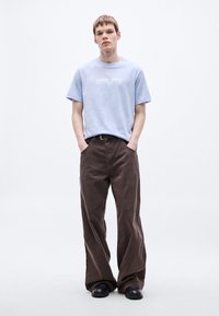 Svetlomodré bavlnené tričko s bielym logom, kombinované s voľnými hnedými kordovými nohavicami a čiernymi koženými topánkami. Jednoduchý, neformálny outfit.