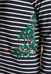 Marineblauwe en witte gestreepte stof met geborduurde groene dennen takken, roze ornamenten en gouden sterren die de vorm van een kerstboom vormen.