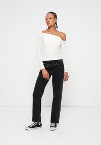 Bela off-shoulder majica s dolgimi rokavi v kombinaciji z visokimi črnimi kavbojkami z srebrnimi okrasnimi studenci ob straneh ter črnimi platnenimi supergami.