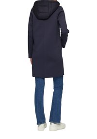 Cappotto lungo blu navy con cappuccio imbottito nero, maniche aderenti e taglio dritto. Indossato su jeans blu e sneakers bianche.