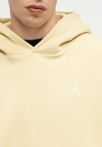 Κοντινό πλάνο ατόμου που φοράει ανοιχτό κίτρινο φούτερ με κουκούλα και λευκό λογότυπο Jumpman στο αριστερό μέρος του στήθους και μαύρο πουκάμισο από κάτω.