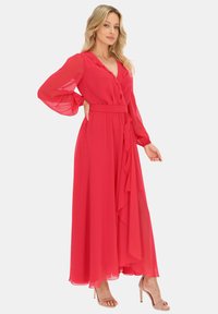 Czerwona maxi sukienka z szyfonu z długimi rękawami, z przodu zapinana na guziki, z falbanami i pasującym paskiem. Noszona z nude sandałami na wysokim obcasie.