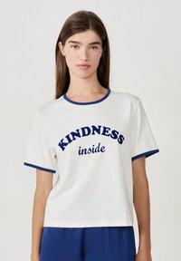 Joven mujer con cabello largo y marrón, vistiendo una camiseta blanca con ribetes azules y las palabras "KINDNESS inside" en texto azul.
