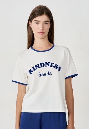 Joven mujer con cabello largo y marrón, vistiendo una camiseta blanca con ribetes azules y las palabras "KINDNESS inside" en texto azul.