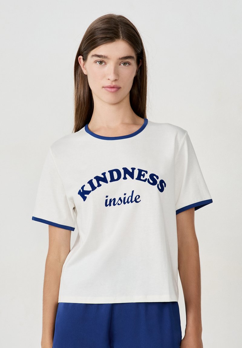 Joven mujer con cabello largo y marrón, vistiendo una camiseta blanca con ribetes azules y las palabras "KINDNESS inside" en texto azul.