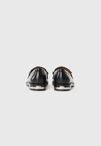 Mocassins en cuir noir avec des accents décoratifs en argent au niveau du talon, montrés de l'arrière sur un fond blanc uni.