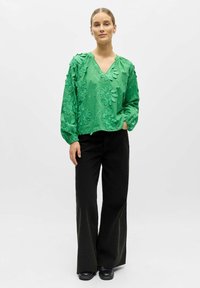 Femme debout portant un chemisier à manches longues vert brodé de fleurs, un pantalon large noir et des chaussures noires, sur un fond clair uni.