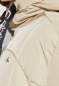 Beige pufferjakke med en blank overflade, der har et quiltet design, sort mærke-snor og logodetalje på stoffet.