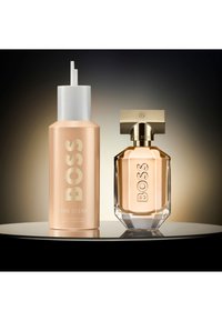 BOSS The Scent parfyme-set med en ferskenfarget refillflaske og en fasettert glassparfymeflaske med gullhatt på en reflekterende overflate.