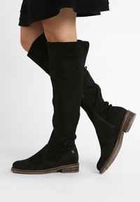 Czarne zamszowe kozaki za kolano z okrągłym noskiem, bocznym zamkiem błyskawicznym oraz teksturowanym, drewnianym obcasem typu blok. Minimalistyczny design z subtelnymi elementami metalowymi.