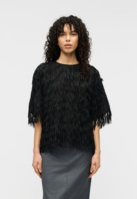 Μαύρο τοπ από υλικό με υφή και φούντες, με φαρδιά κοντά μανίκια, σε συνδυασμό με εφαρμοστή γκρι φούστα, παρουσιάζεται σε ουδέτερο φόντο.