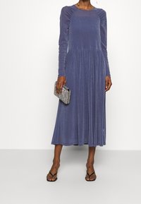Midikappa violetti pitkähihainen mekko, jossa on laskostettu helma ja teksturoitu kangas. Yhdistetty hopeiseen clutch-laukkuun ja mustiin sandaaleihin.