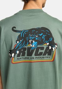 Groene katoenen t-shirt met een gedurfde zwarte panterafbeelding, blauwe accenten en de tekst "RVCA NATUUR TEGEN INDUSTRIE" in grote letters.
